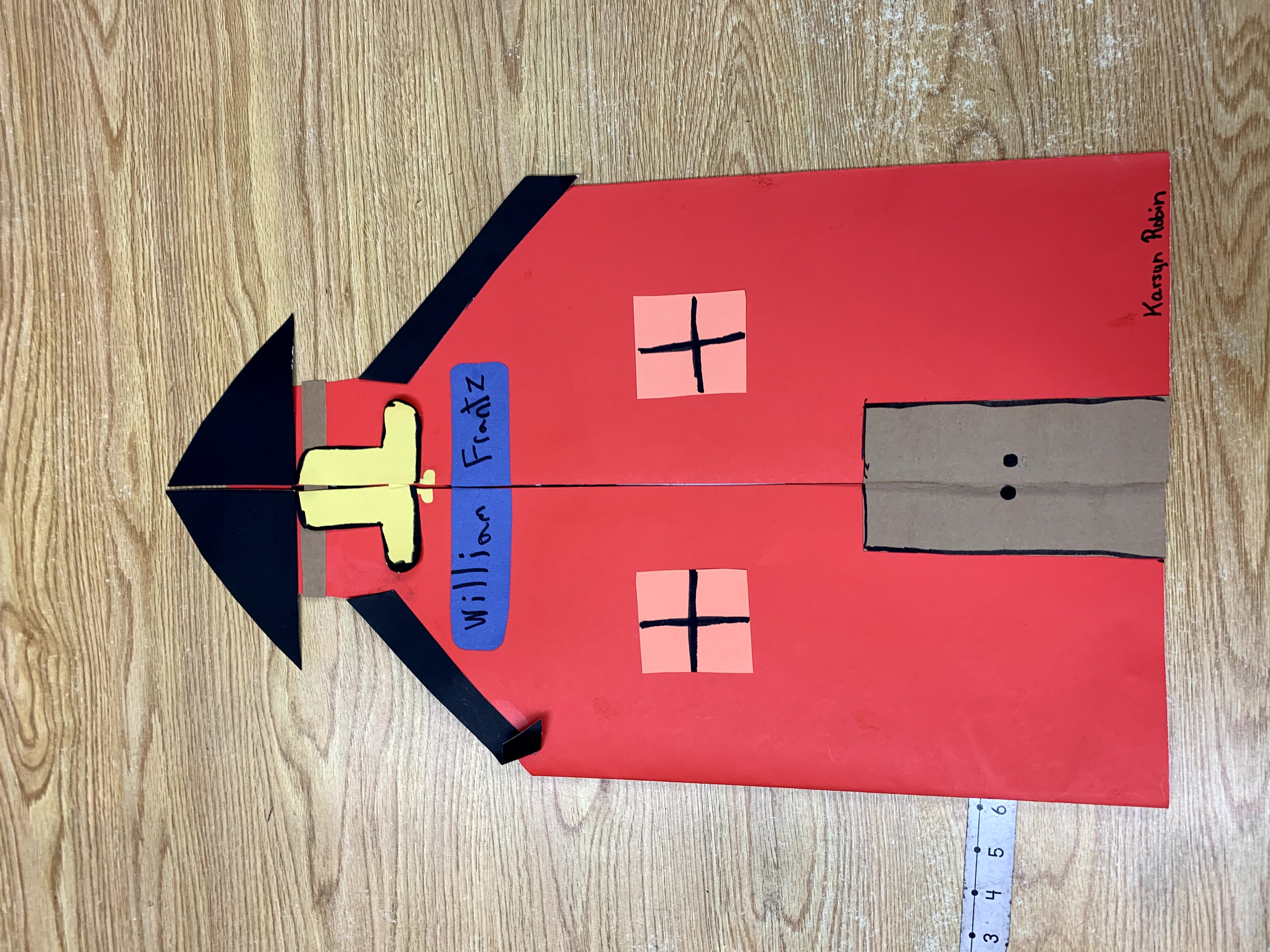 A red paper schoolhouse craft with a bell and "William Friedz" written on it sits on a wooden surface with a ruler to its left.