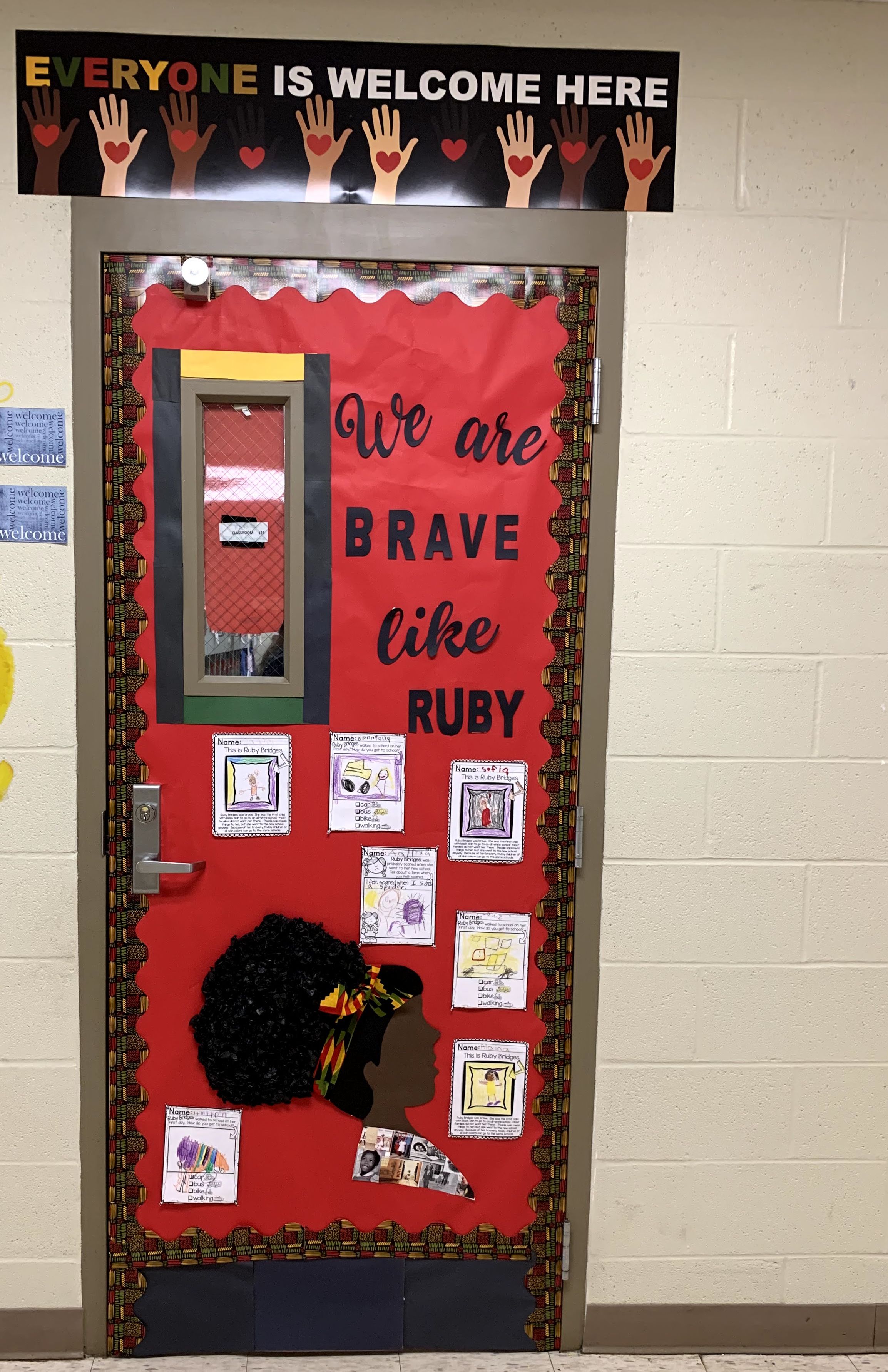 A classroom door decorated with a silhouette of a Black woman and children's artwork is framed by a colorful border, while a banner above reads "EVERYONE IS WELCOME HERE" against a beige wall.