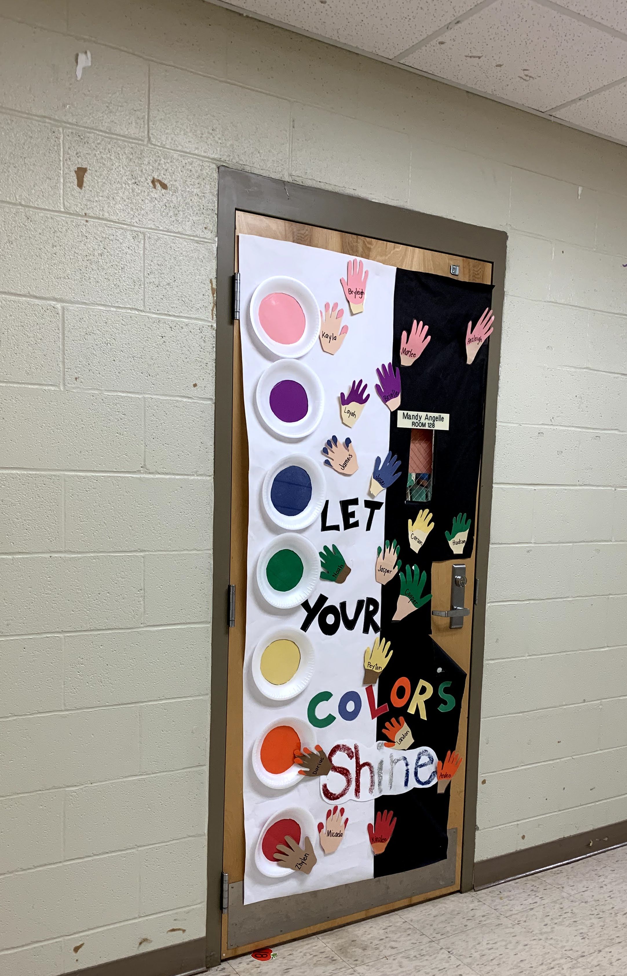 Colorful handprints and paint palettes adorn a classroom door, set against a white brick hallway.