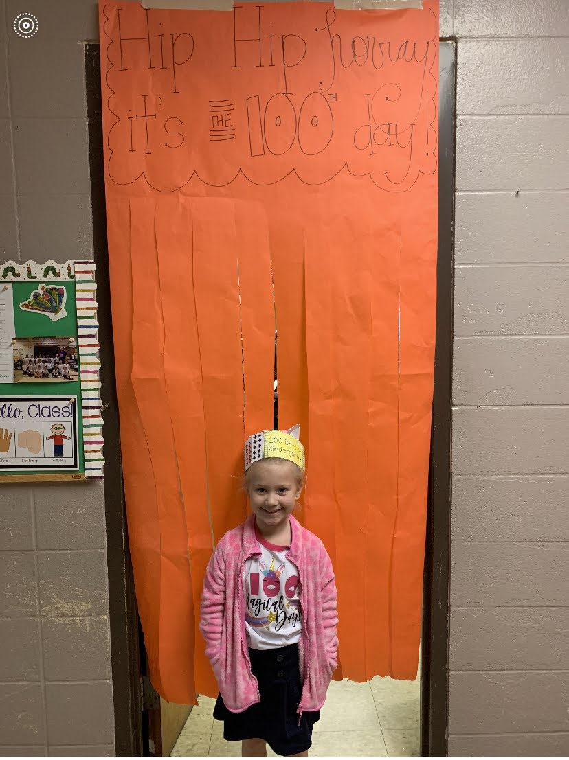 A young girl in a pink cardigan and a festive headband stands in front of an orange-papered doorway decorated with a "Hip Hip Hooray! It's the 100th day!" banner, while a bulletin board with colorful student work is visible to the left.