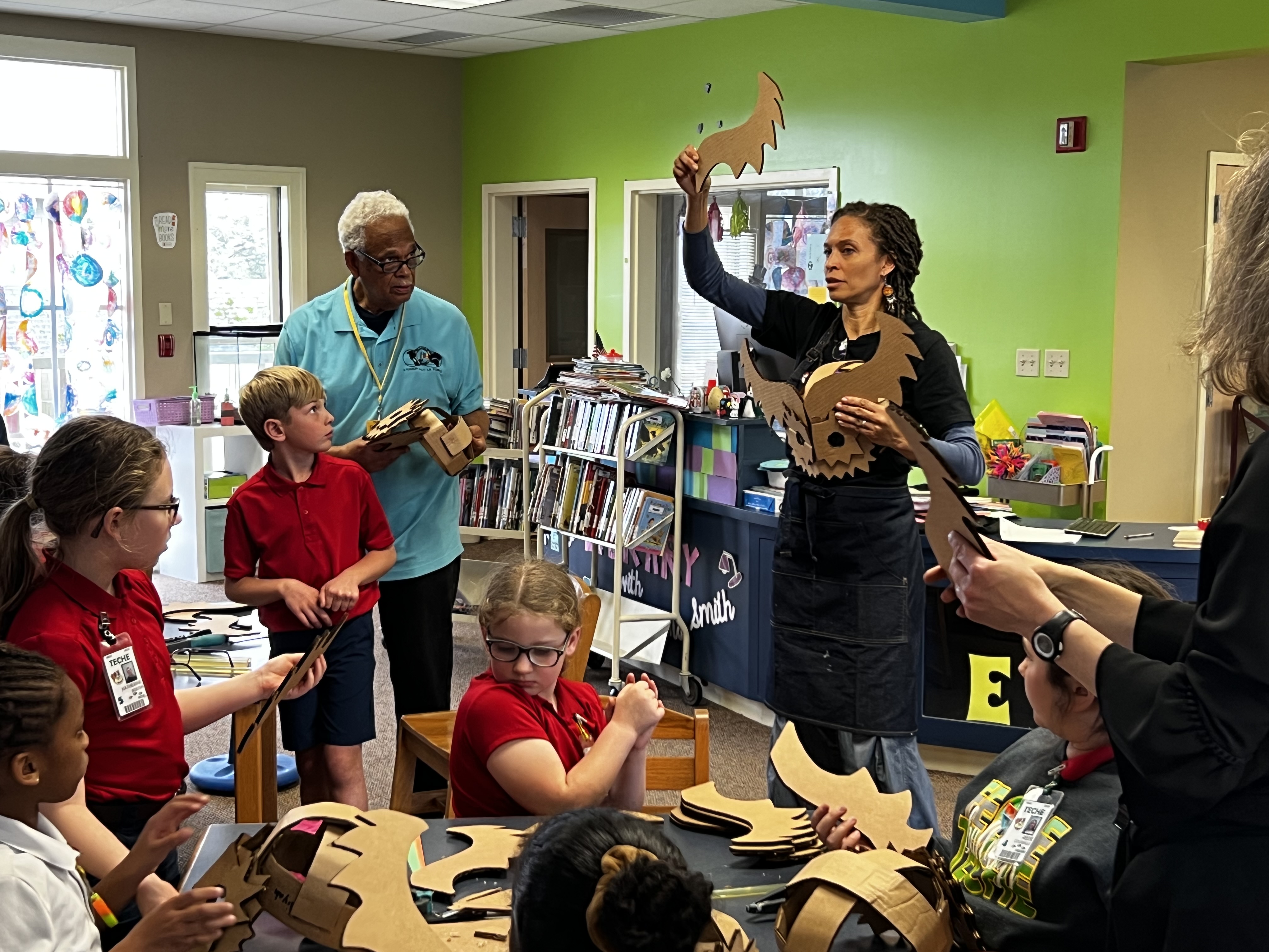 In the foreground, children in red shirts are gathered around a table, crafting with cardboard, while in the background, two adults, one in a blue shirt and the other in black, lead the activity in a brightly lit classroom with bookshelves.