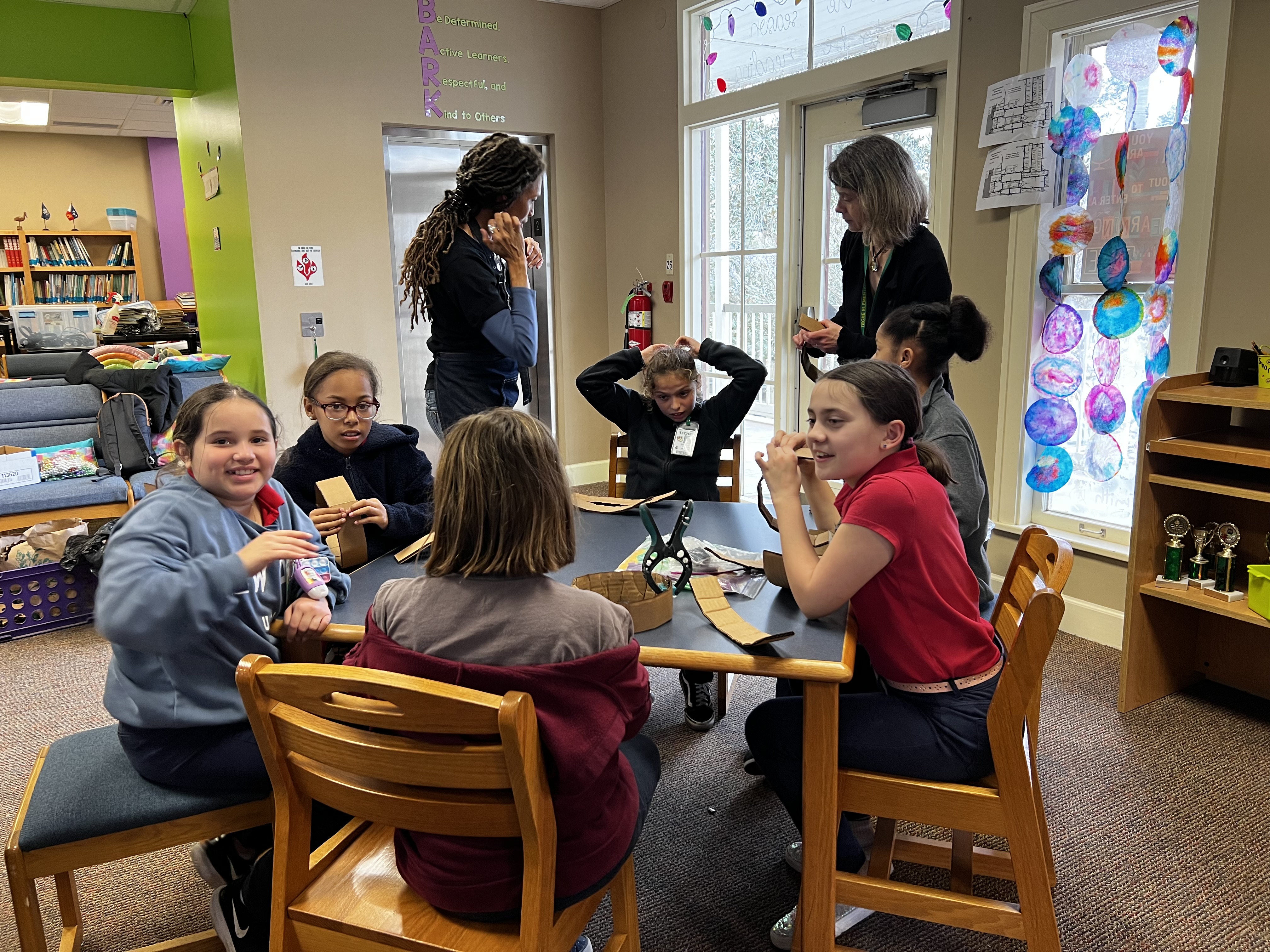 A group of young girls are gathered around a table in a classroom, with bookshelves and colorful decorations visible in the background.