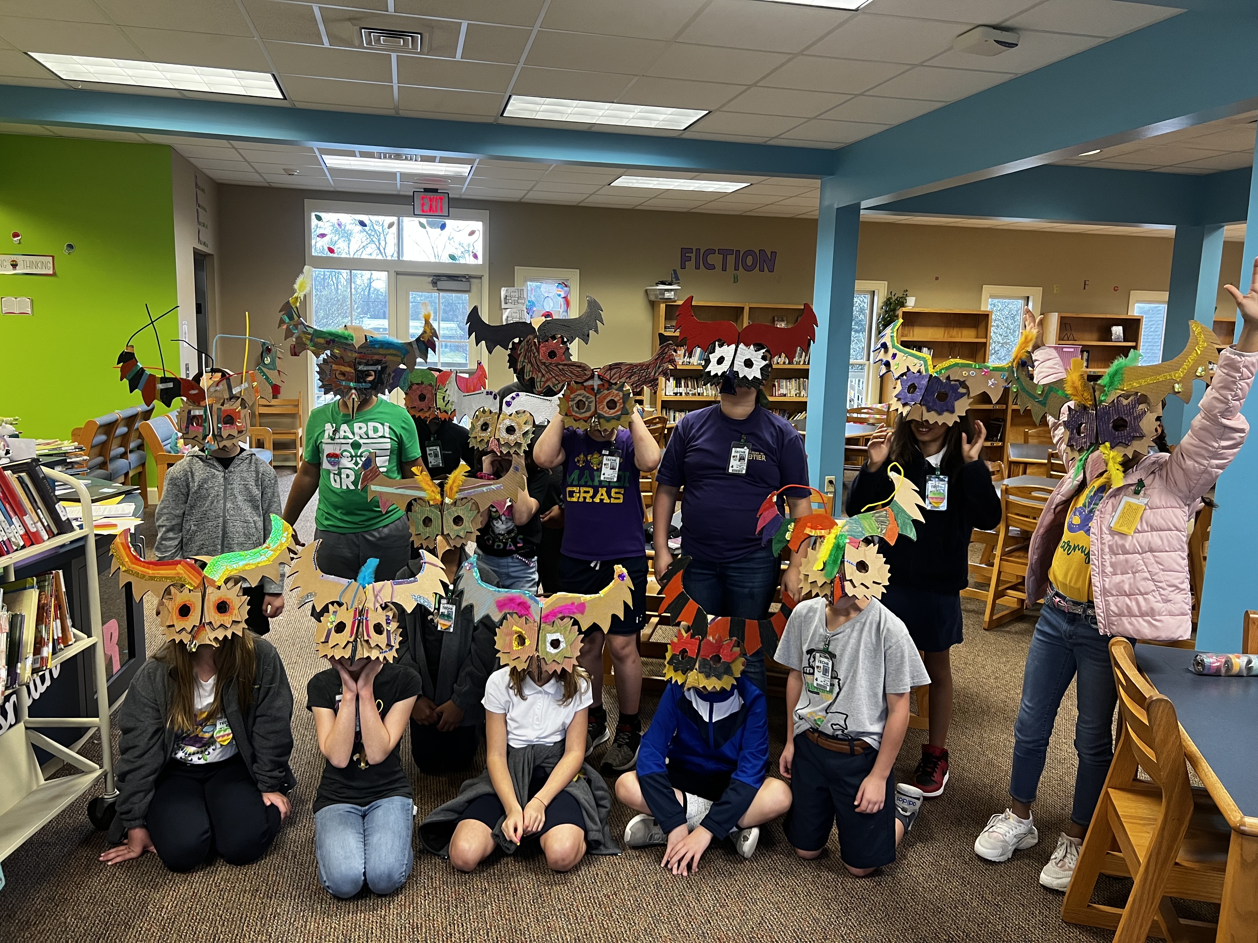 A group of children and adults wearing colorful, handmade owl masks pose in a library with bookshelves and a "Fiction" sign in the background.
