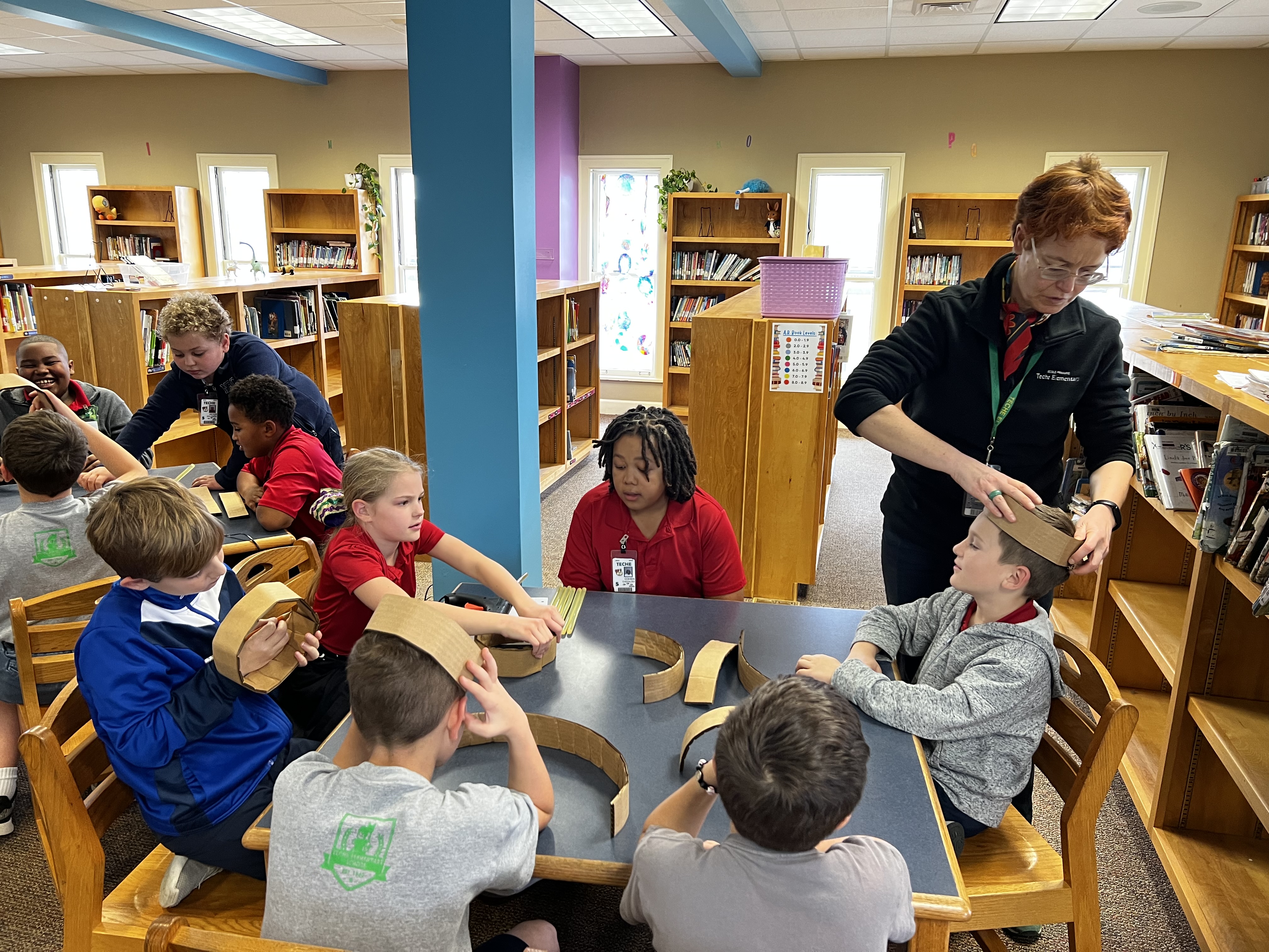 Children in the foreground are working on a craft project at tables in a library, while a teacher assists them amidst bookshelves in the background.