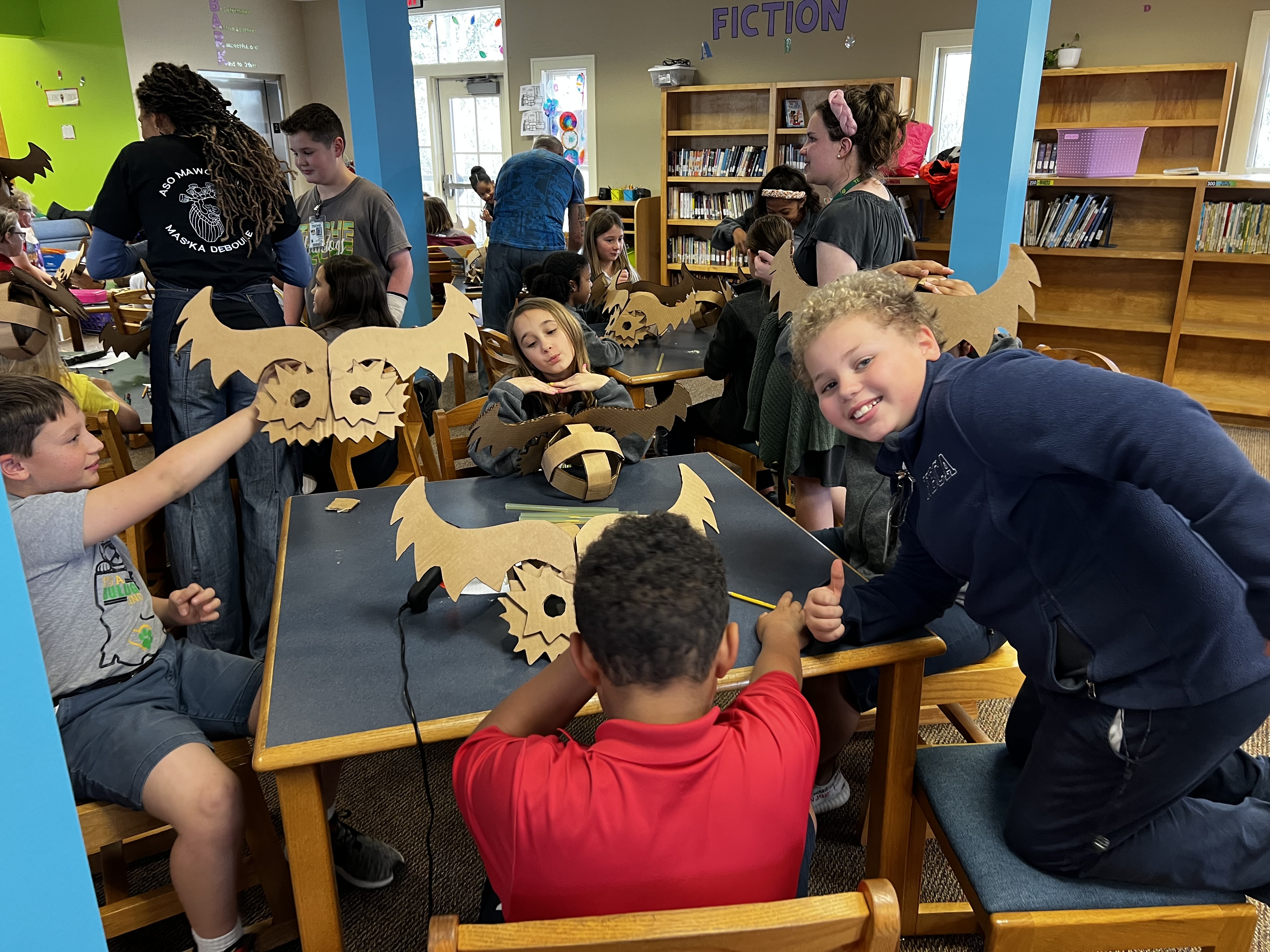 Children in the foreground are working on crafts at tables, while in the background, bookshelves filled with books line the walls of a library.