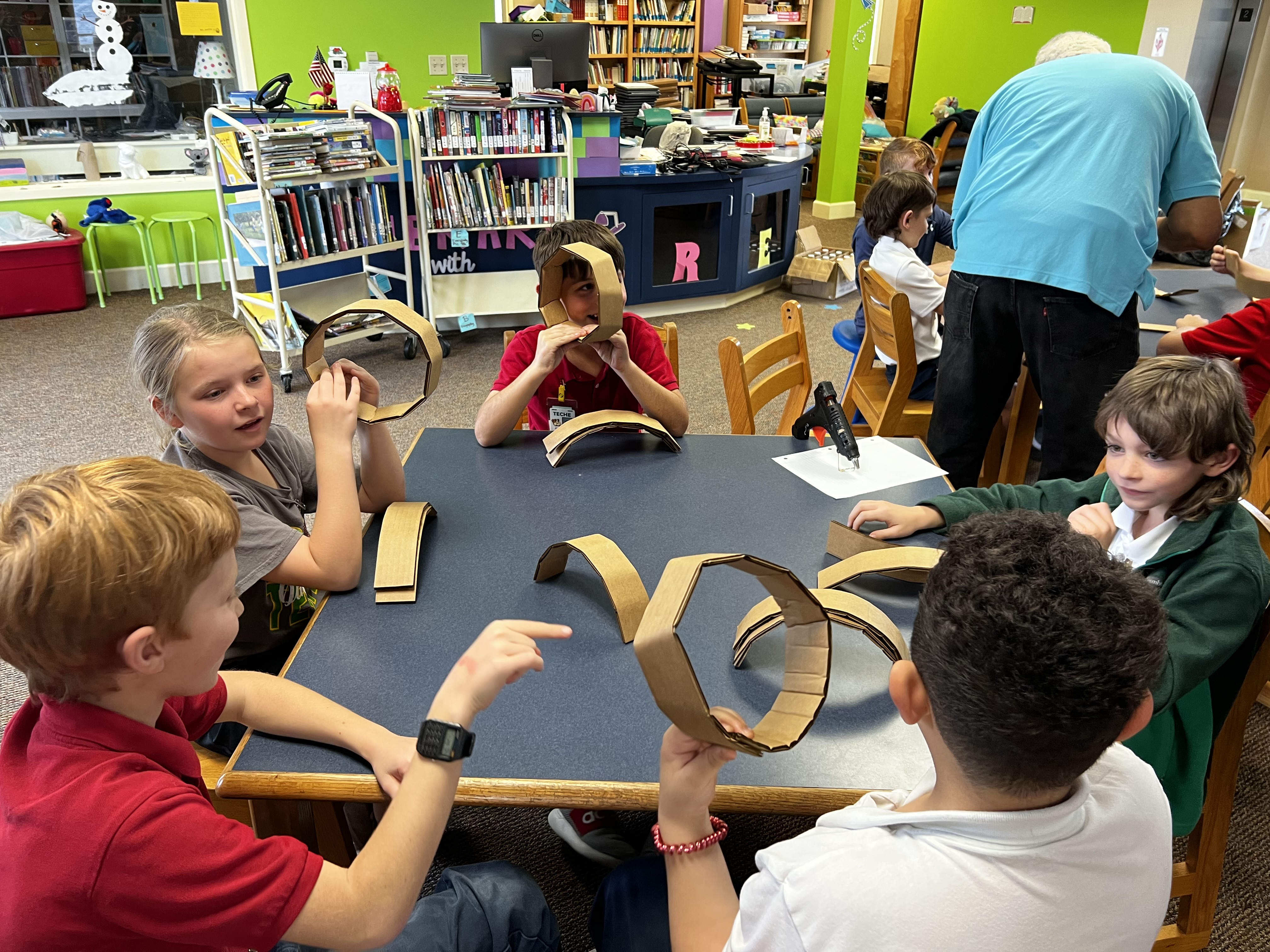 Children in the foreground are engaged in a craft project with cardboard, while in the background, a library setting with bookshelves and other students is visible.