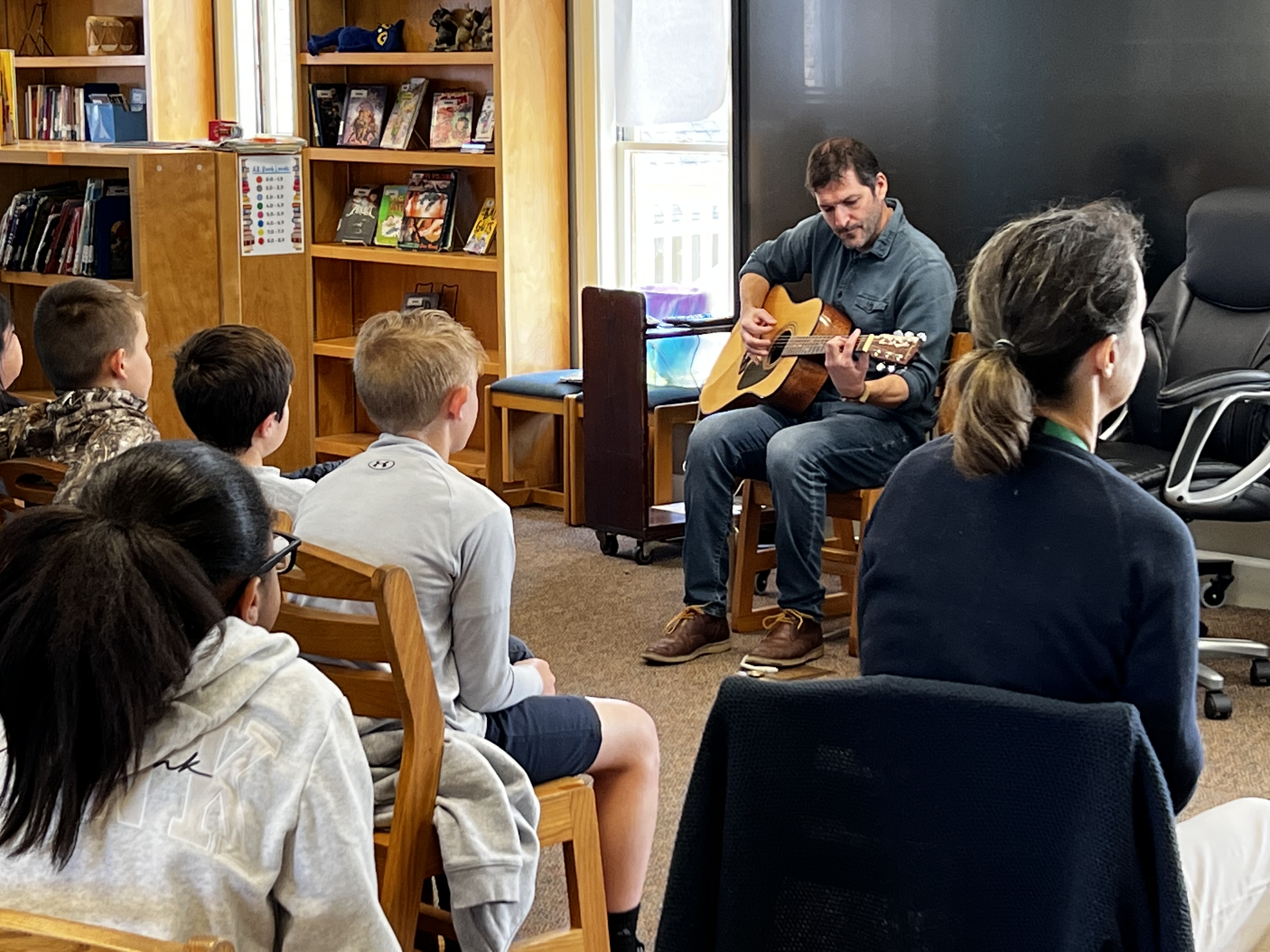 A man plays guitar for a group of children sitting in chairs in front of bookshelves.