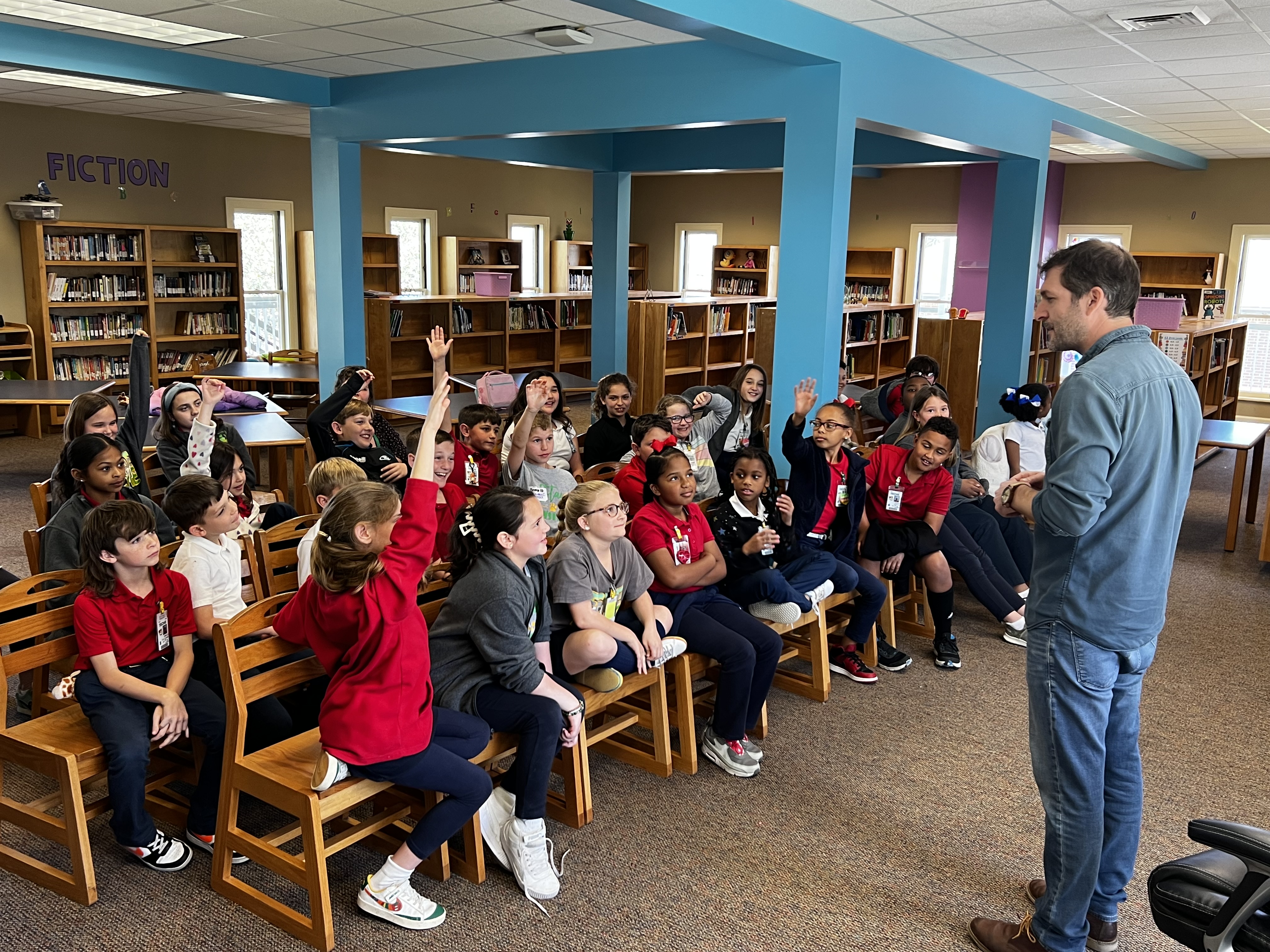 A man in jeans and a blue shirt stands facing a group of children in a library, where bookshelves filled with books line the walls.