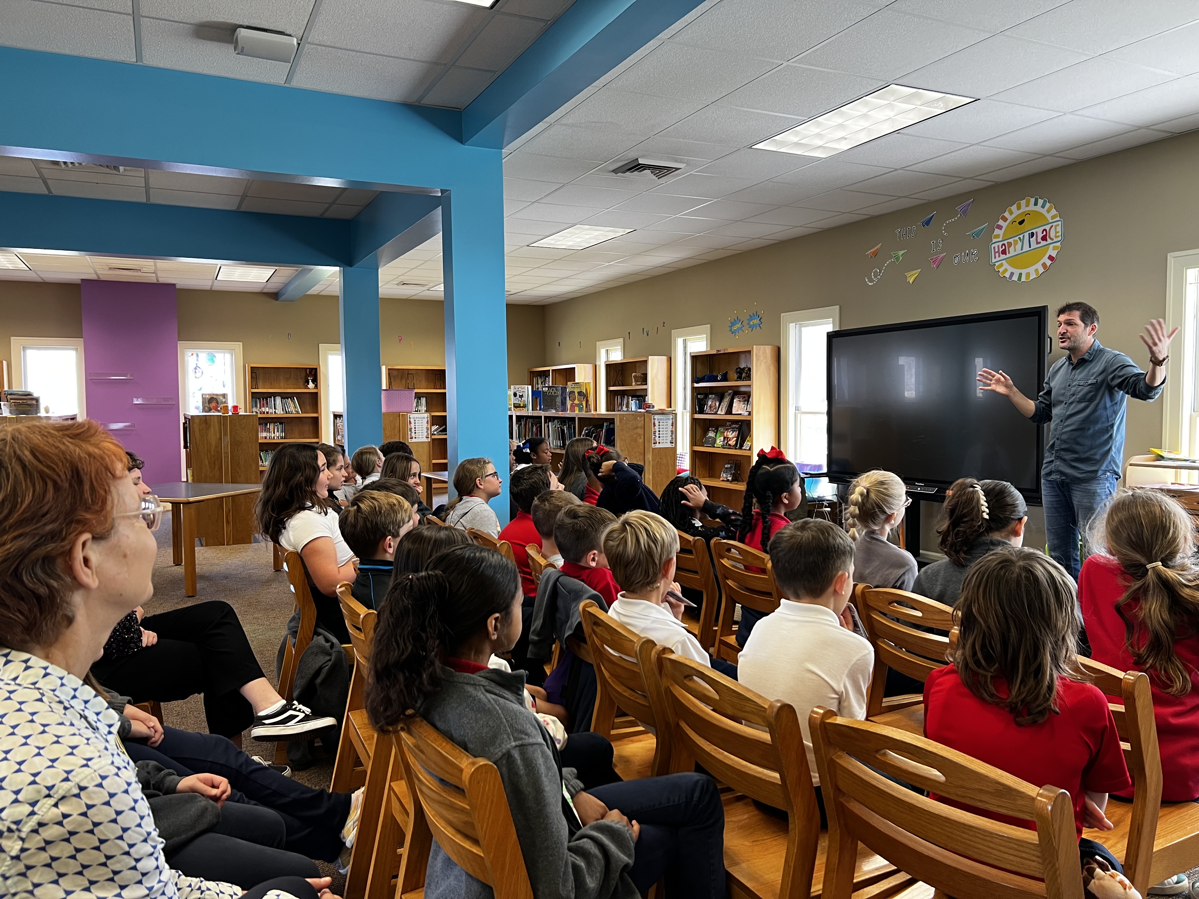 A teacher presents to a classroom of students seated in wooden chairs, with bookshelves lining the background walls.