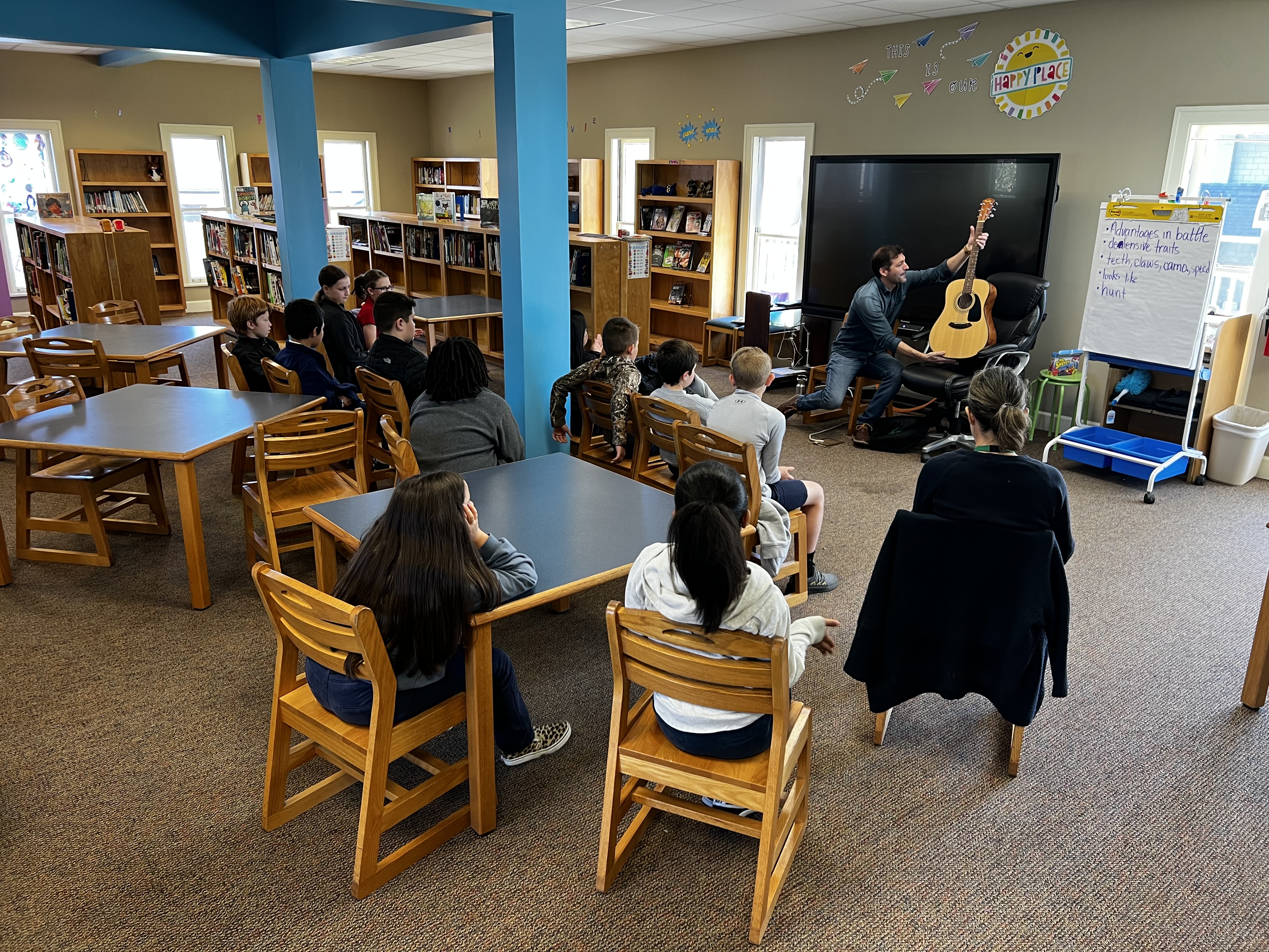 Children sit at tables in the foreground, facing a man playing a guitar in front of a whiteboard in the background, with bookshelves lining the walls.