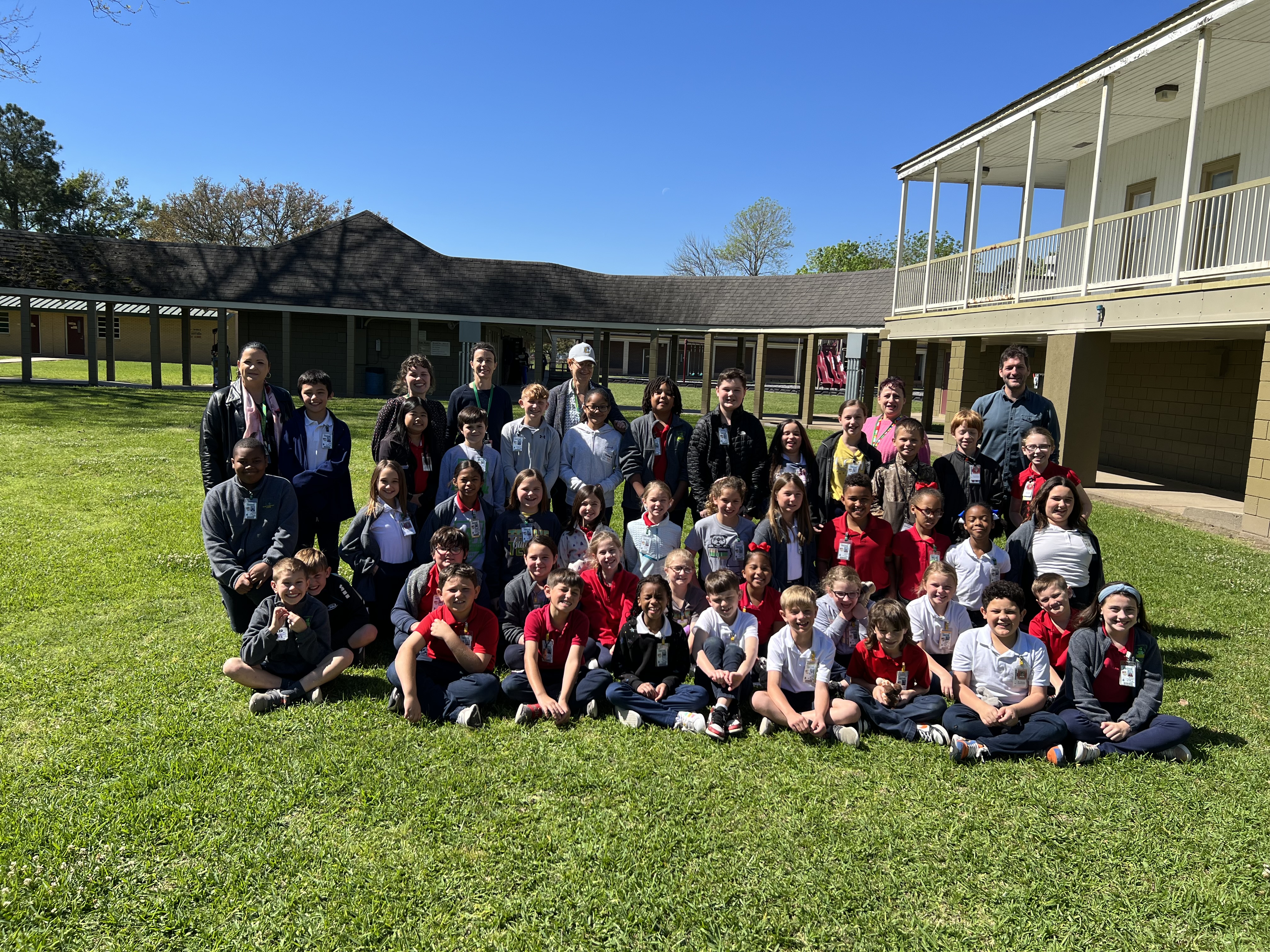 A large group of children and adults pose for a photo on a grassy lawn in front of a school building with a covered walkway.