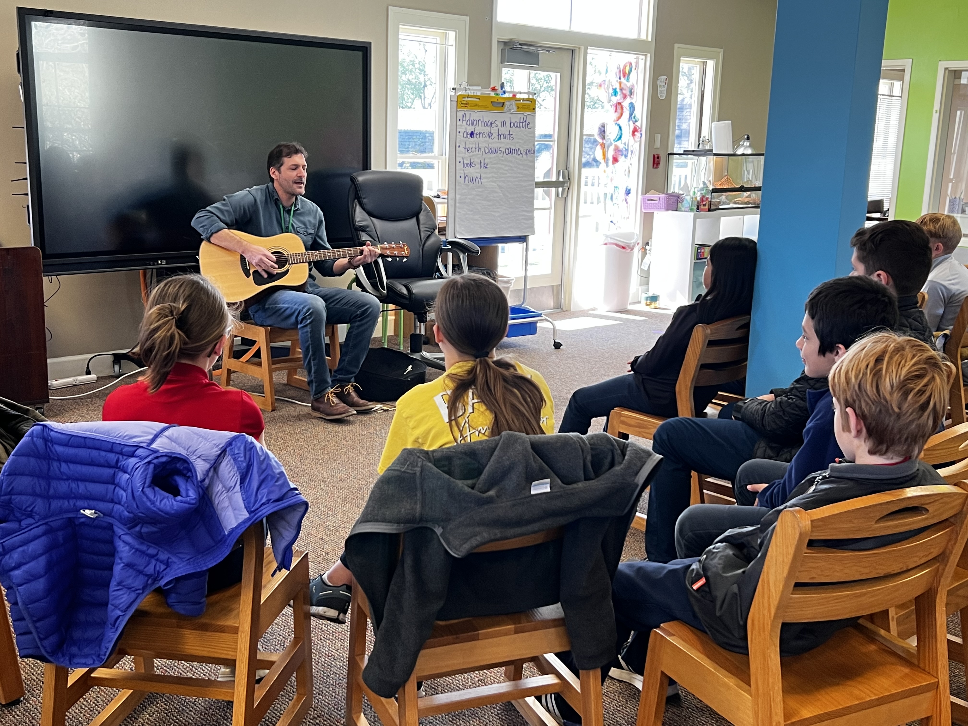 A man plays guitar for a group of children sitting on chairs in a brightly lit room with a large screen and whiteboard in the background.