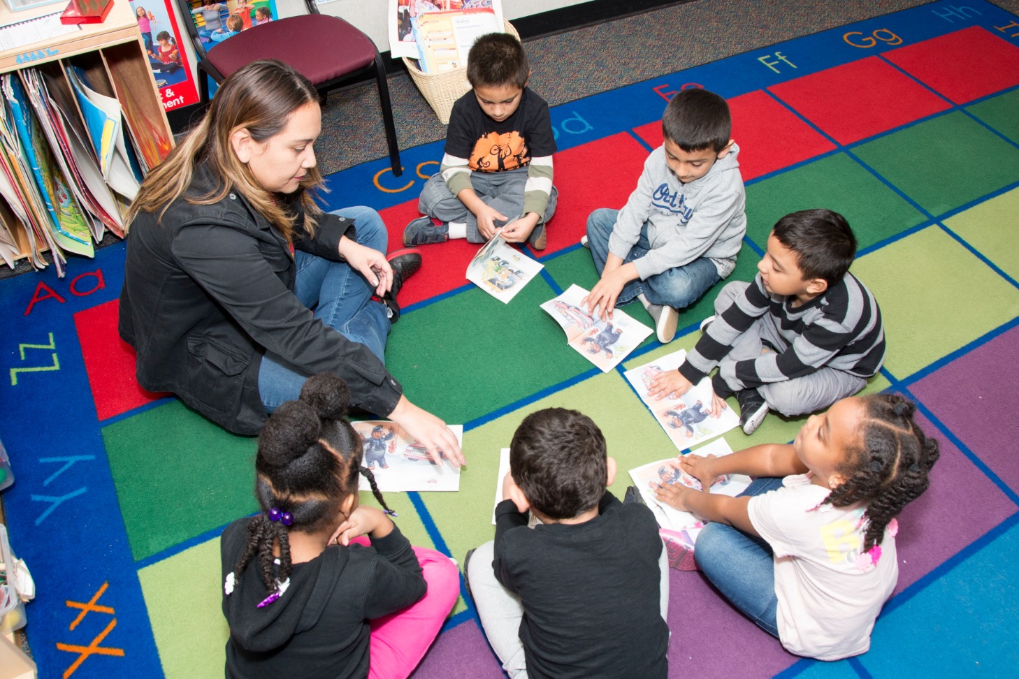 A group of children sitting on a colorful mat, with a teacher reading.
