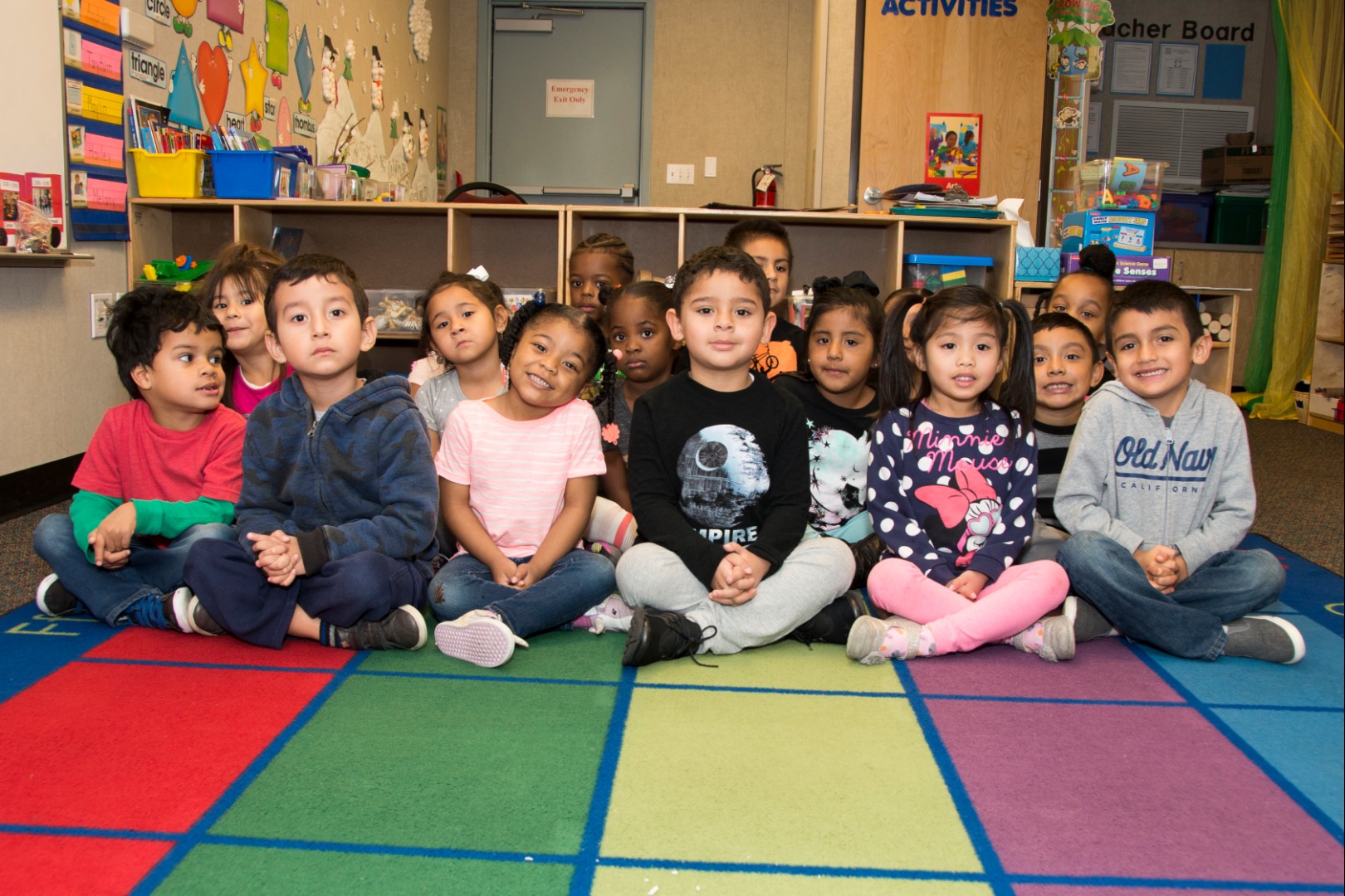 Children sitting on a colorful carpet in their classroom with displays visible in the background.