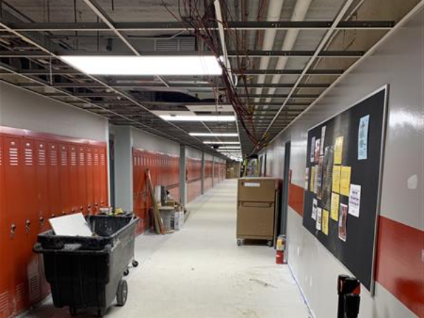 A long, empty school hallway with orange lockers on the left and a bulletin board on the right, leading to a dimly lit background with exposed ceiling pipes.