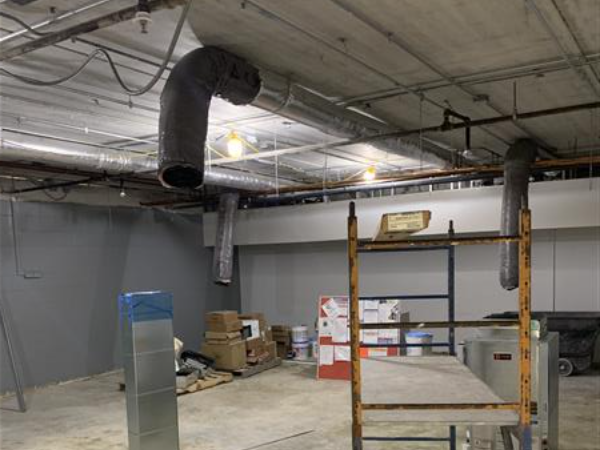 In the foreground, a metal duct and scaffolding are visible, while the background reveals a room with exposed pipes and boxes.