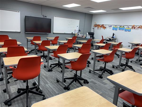Rows of red chairs and light-colored desks fill the foreground of a modern classroom, while whiteboards and a large screen adorn the gray walls in the background.