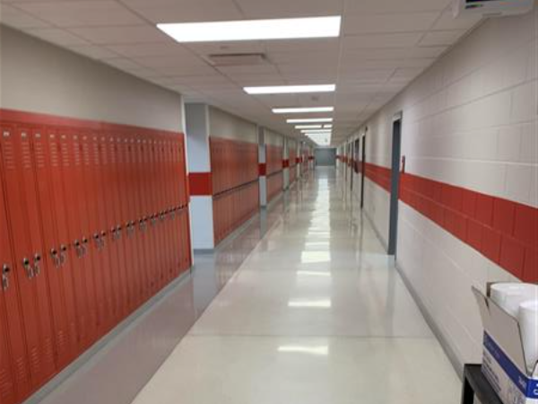 A long, brightly lit school hallway with rows of orange lockers on the left and white walls with a red stripe on the right stretches into the distance.