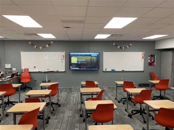 In the foreground, rows of desks with orange chairs face a large screen, while the background features whiteboards and a gray wall.