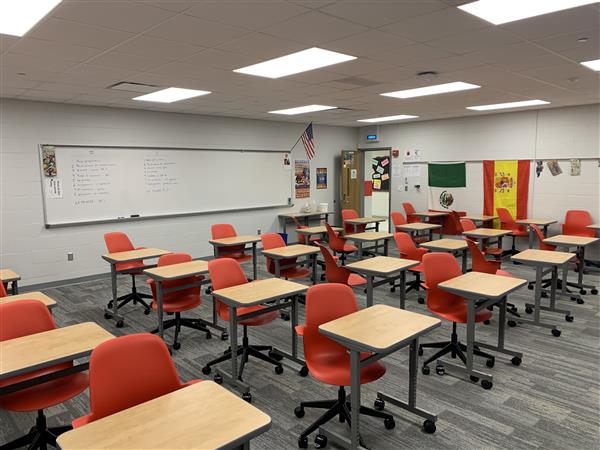 Rows of empty desks with orange chairs fill the foreground of a classroom, while a whiteboard with writing and flags hang on the wall in the background.