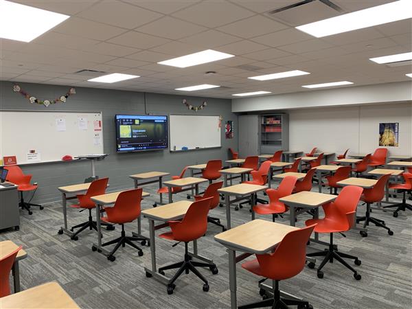 Rows of empty red chairs and light-colored desks fill the foreground of a modern classroom, while a large screen and whiteboards adorn the gray wall in the background.