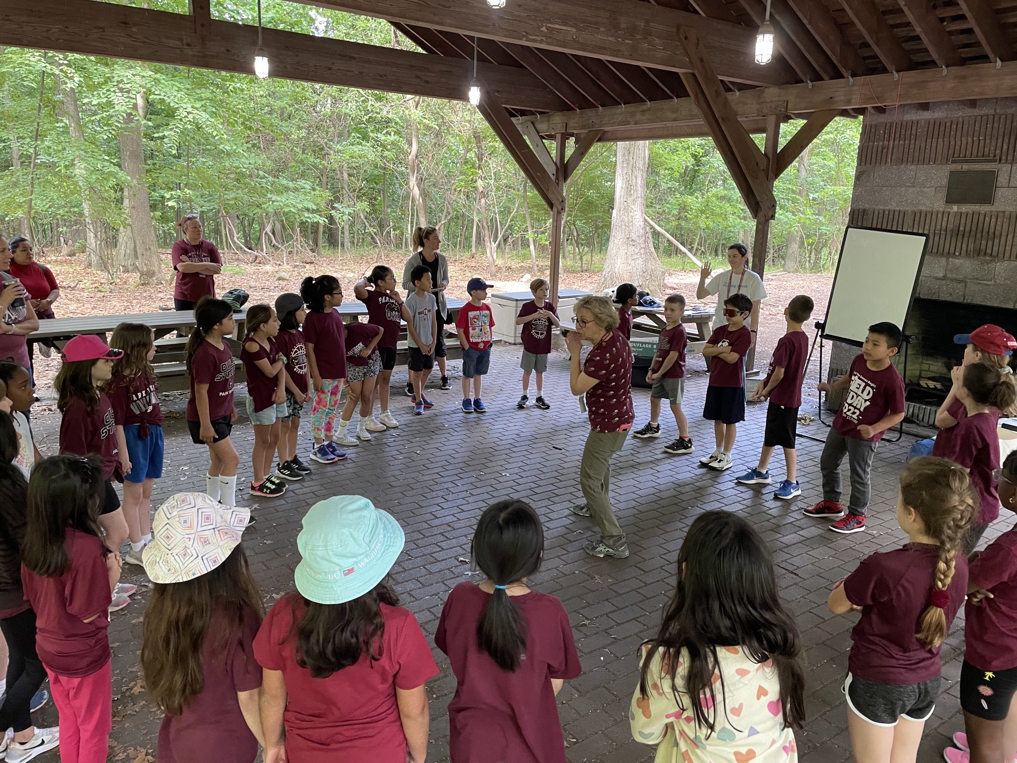 Children in maroon shirts stand in a circle under a wooden pavilion, with trees and a whiteboard visible in the background.