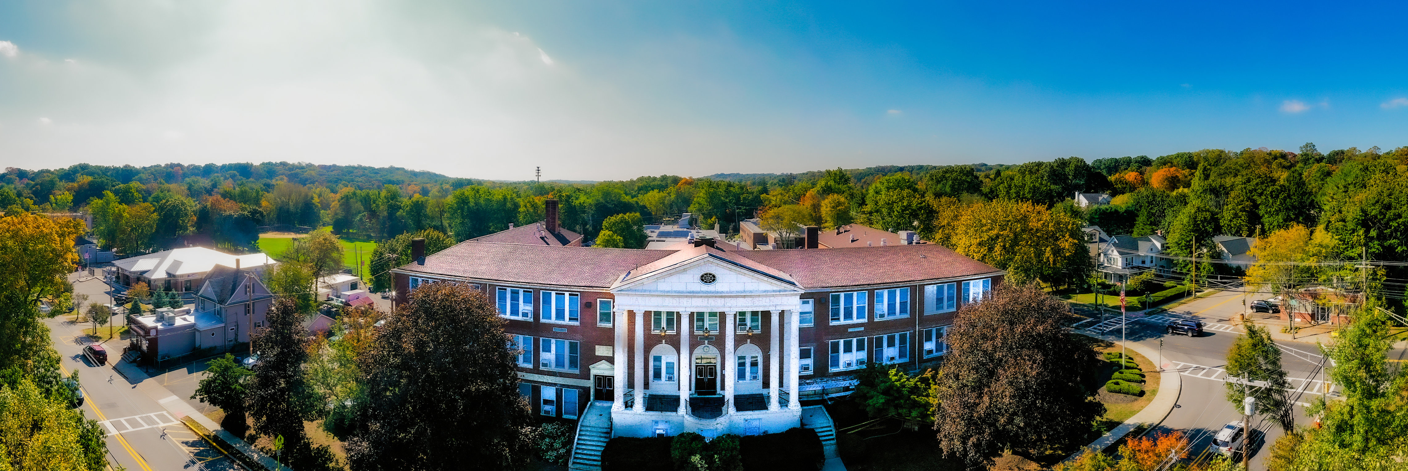 A grand brick building with white columns stands in the foreground, surrounded by lush green trees and a clear blue sky in the background.