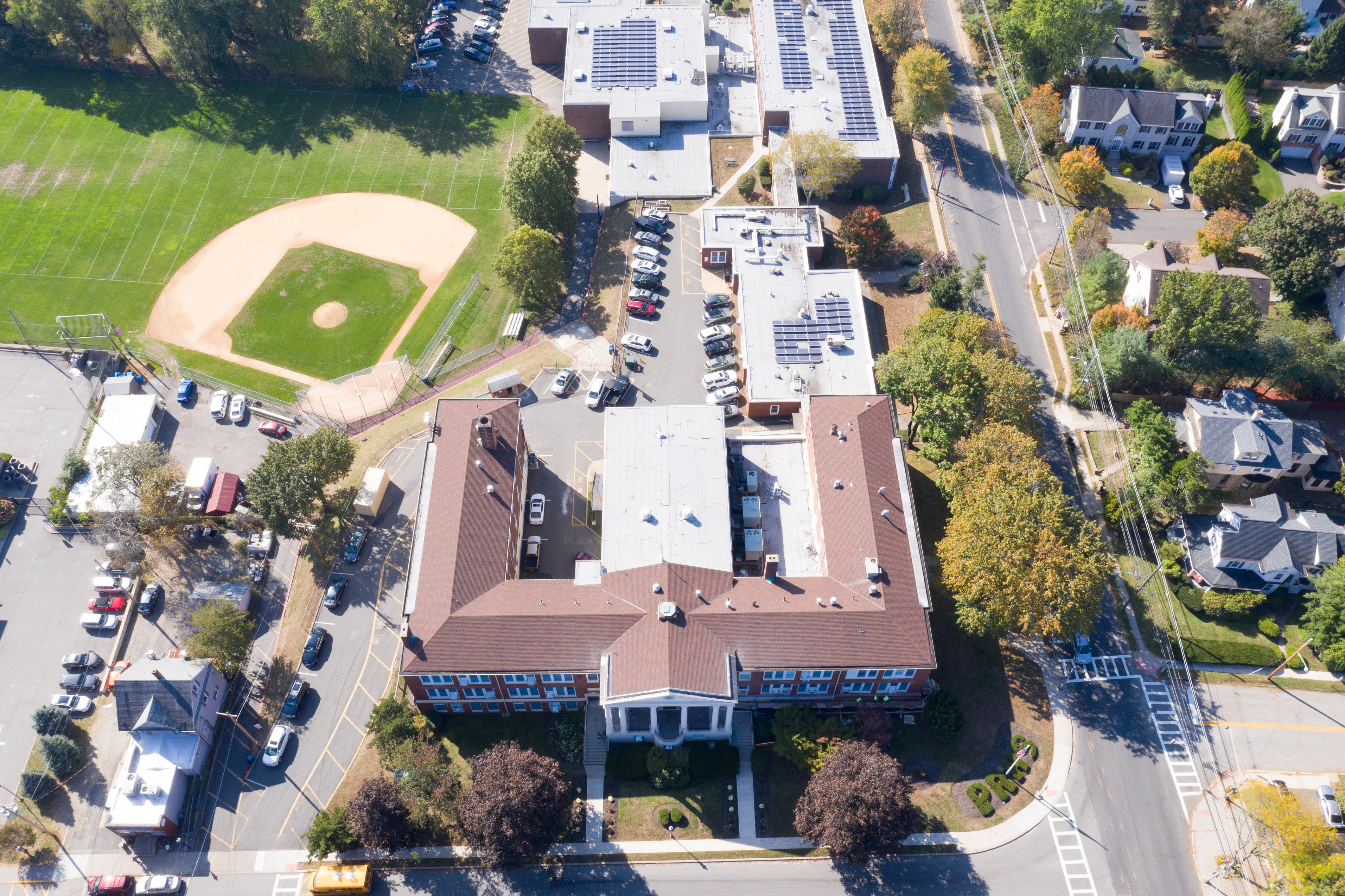 An aerial view captures a U-shaped school building with a baseball field to its left, surrounded by trees and residential homes.