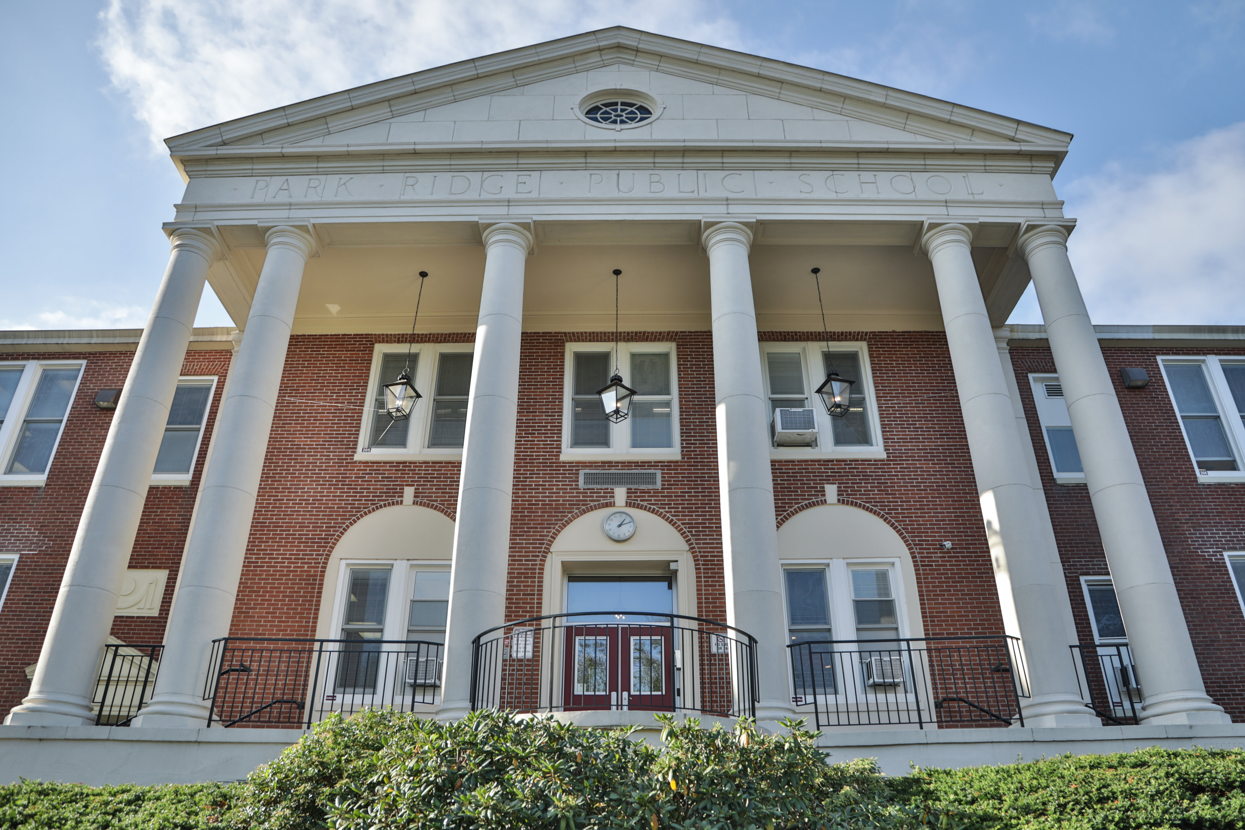 A grand brick building with large white columns and a pediment stands behind lush green bushes in the foreground, under a clear blue sky.