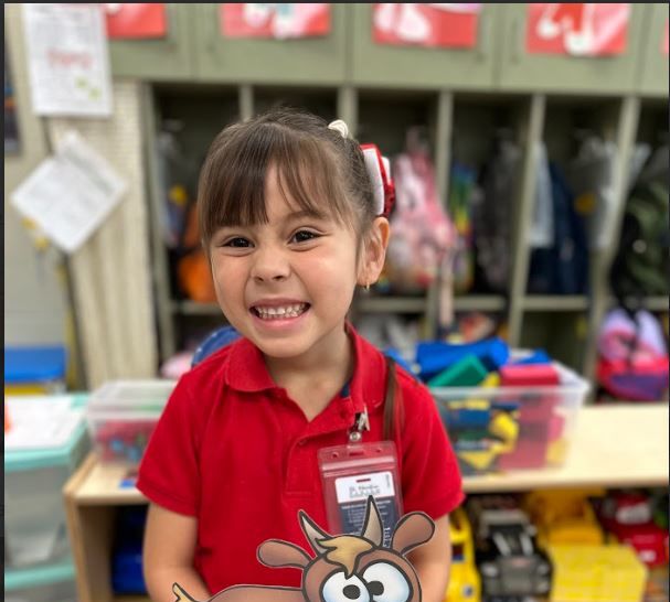 A smiling young girl in a red shirt holds a cutout of a goat in front of a classroom background with cubbies and supplies.