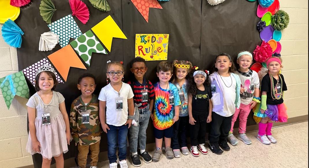 A group of smiling children in colorful outfits stand in front of a festive classroom bulletin board decorated with paper fans and banners.