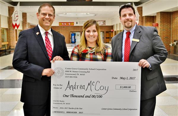 Three people stand in a hallway holding a large novelty check, with a brick wall and school signage in the background.