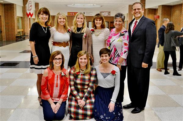 A group of nine people, eight women and one man, pose for a photo in a hallway with a red and white banner visible in the background.