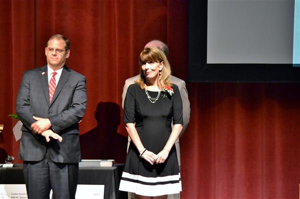 A man in a suit and a woman in a black dress stand on a stage with a red curtain backdrop and a projection screen.