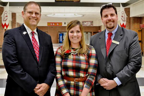 Three smiling individuals in formal attire stand in the foreground, with a school hallway featuring banners and lockers visible in the background.