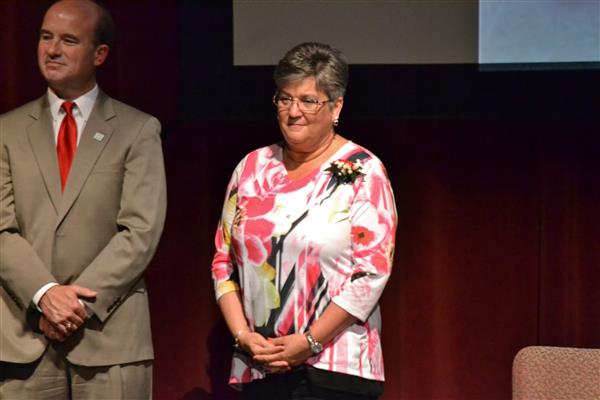 A woman in a floral shirt stands in the foreground with a man in a suit to her left, against a dark red curtain background.