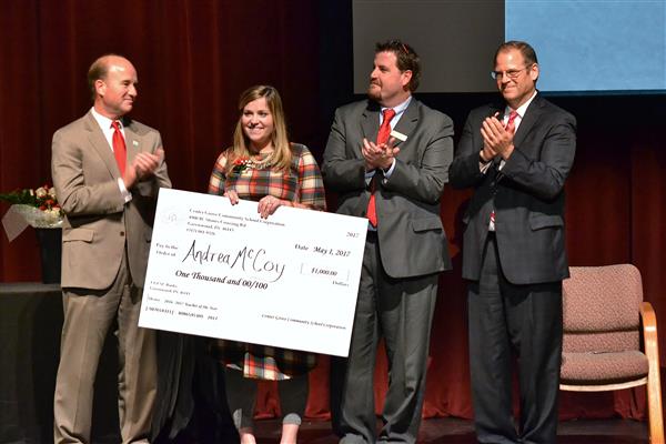 Four people stand on a stage in front of a red curtain, with three men in suits clapping and a woman holding a large check.