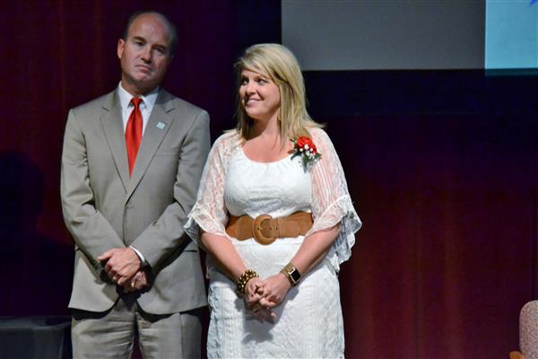 A man in a tan suit and red tie stands beside a woman in a white dress with a brown belt, against a dark red backdrop.