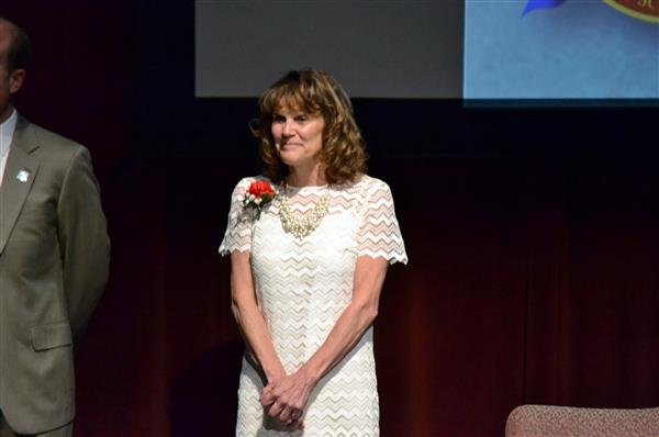 A woman in a white lace dress stands on a stage with a man in a suit to her left, while a dark curtain and a blue banner form the background.