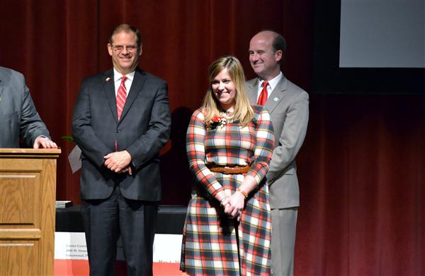 Three people in formal attire stand on a stage with a red curtain backdrop, while a wooden podium is visible in the foreground.