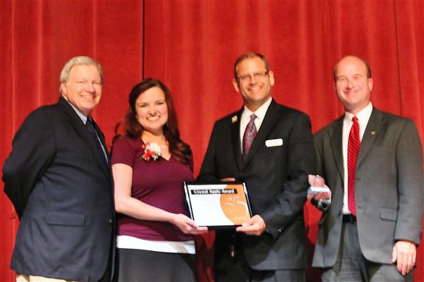 Four people stand in front of a red curtain, with two men on the left and two men on the right, and a woman in the center holding an award.