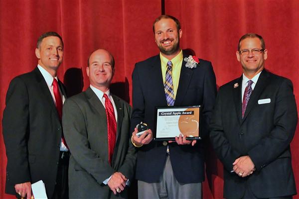 Four men in suits stand in front of a red curtain, with the man in the center holding an award and a small apple.