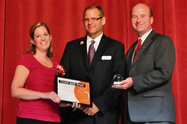 A woman in a pink shirt and two men in suits stand in front of a red curtain, with one man presenting an award to the woman and the other holding a small trophy.