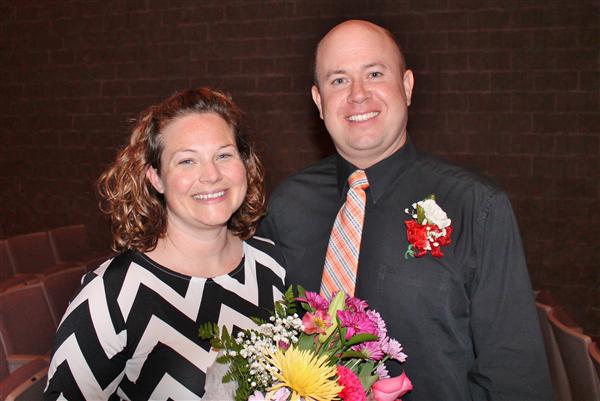 A smiling couple poses with a bouquet of flowers in front of a brick wall and empty theater seats.