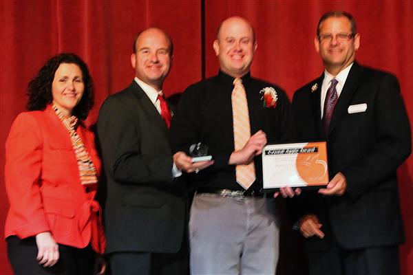 Four people stand on a stage with a red curtain in the background, while two men in the foreground hold an award and a certificate.