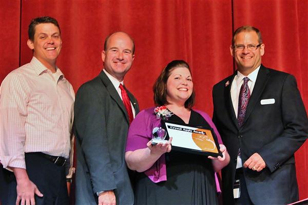 Four smiling people stand on a stage with a red curtain in the background, while the woman in the center holds an award.