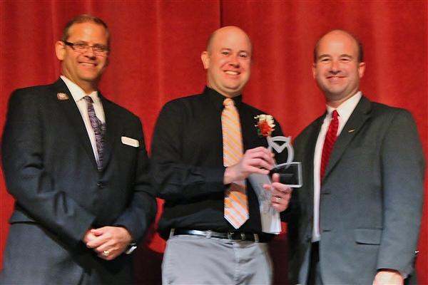 Three men in suits stand on a stage with a red curtain in the background, while the man in the middle holds an award.
