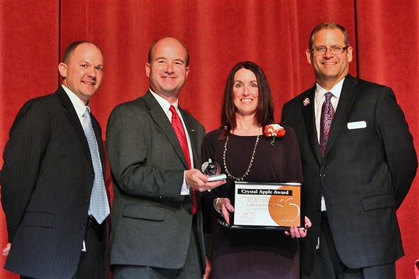 Four people stand in front of a red curtain, with two men on the left and two on the right, holding an award and a certificate.