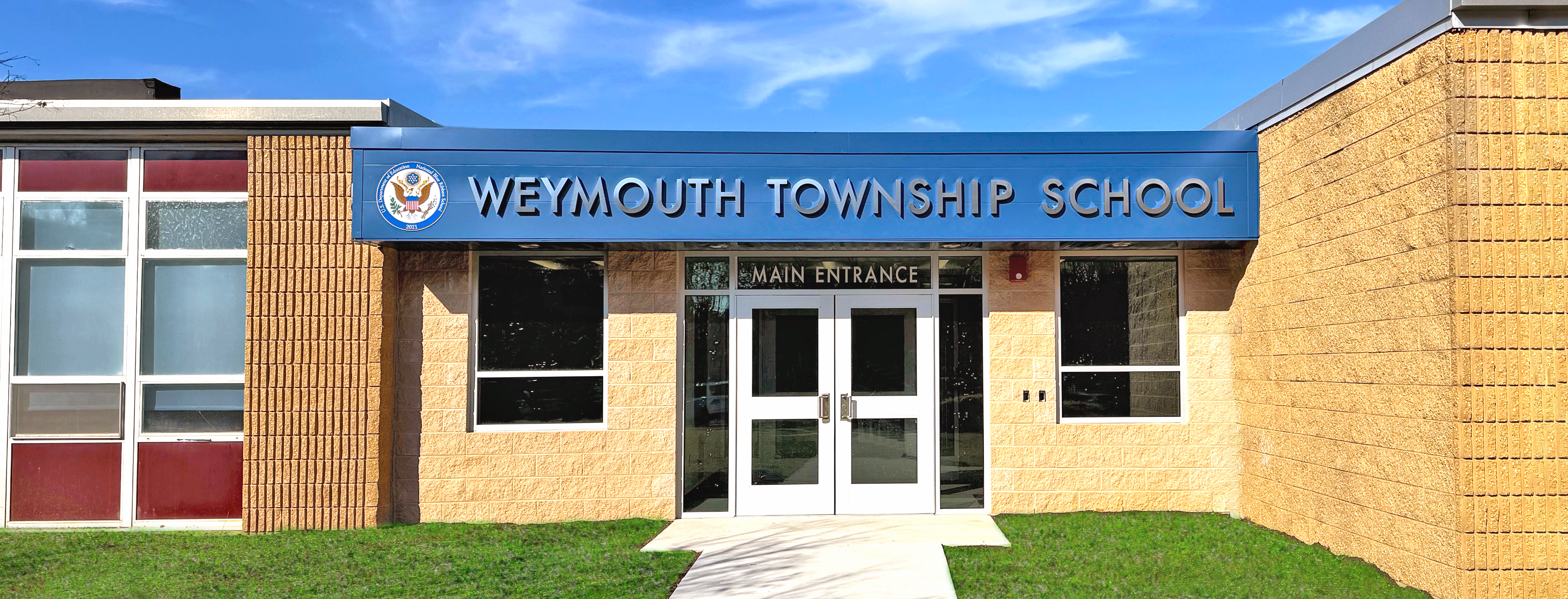 A bright blue sign above the main entrance of Weymouth Township School is set against a tan brick building, with a clear blue sky and white clouds in the background.
