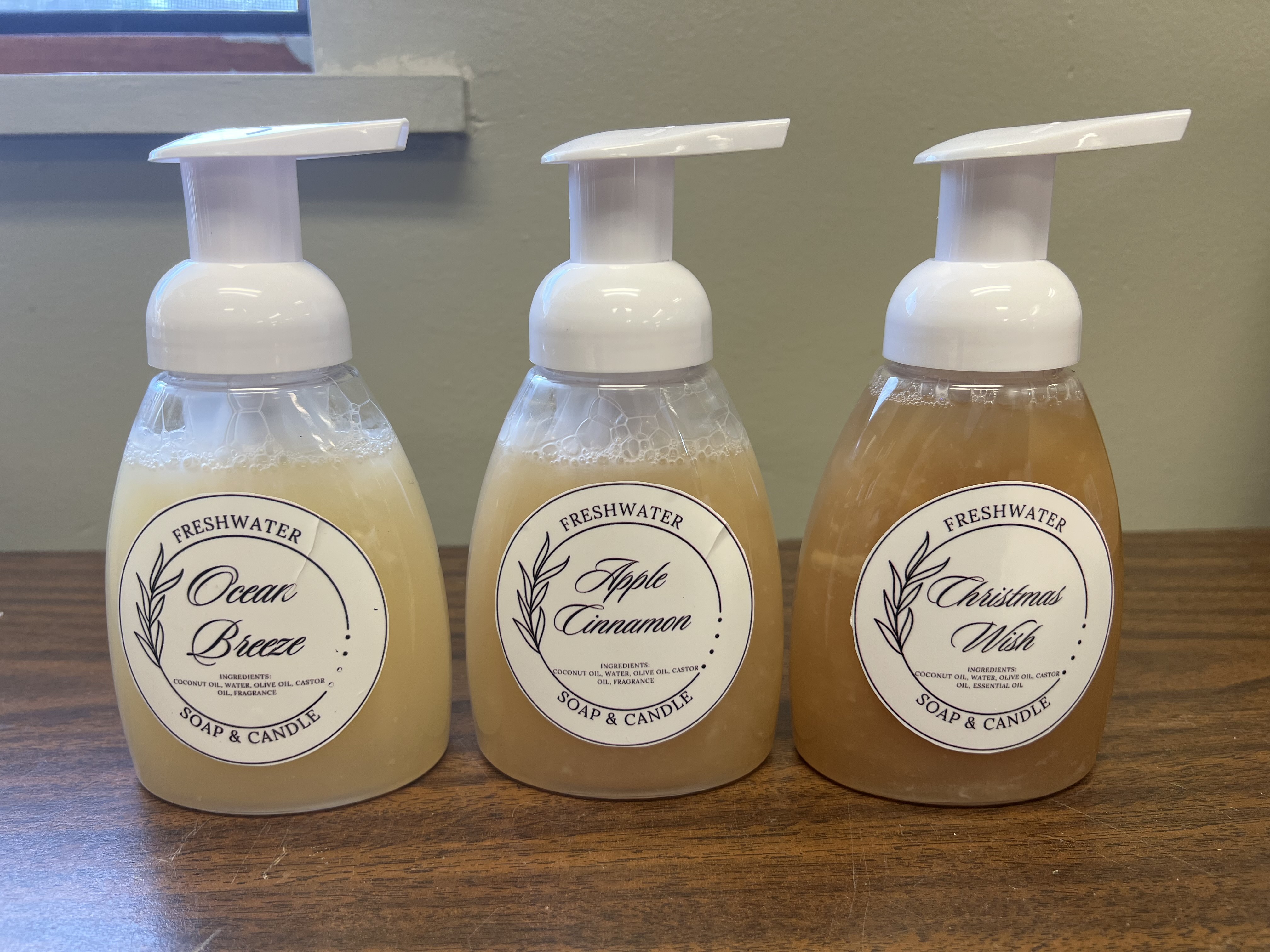 Three bottles of Freshwater soap and candle products, including scents like Citrus Breeze, Apple Cinnamon, and Christmas Mint, are displayed on a wooden surface against a neutral background.